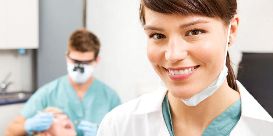 背景中，一名女牙科助理微笑着看着一名牙医正在给病人治疗牙齿