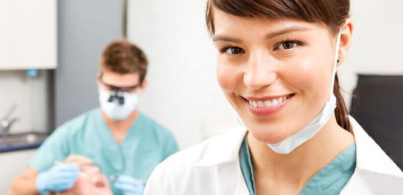 一名女性牙科助理微笑着，背景是一名男性牙科助理在给病人做手术