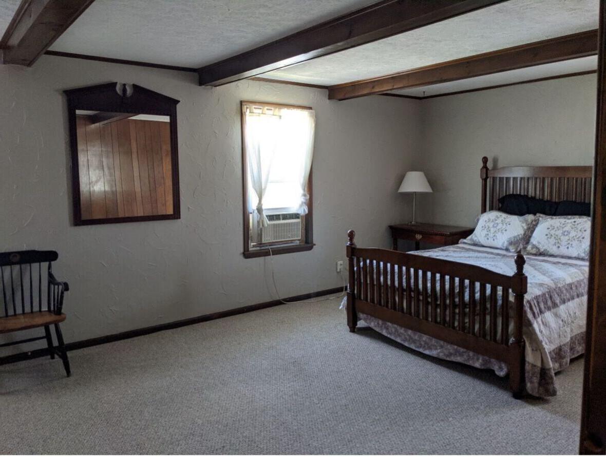 校友之家的一间卧室，有一张木制的大床, 带空调的窗户, nightstand with lamp, wood-framed mirror, and wooden chair.