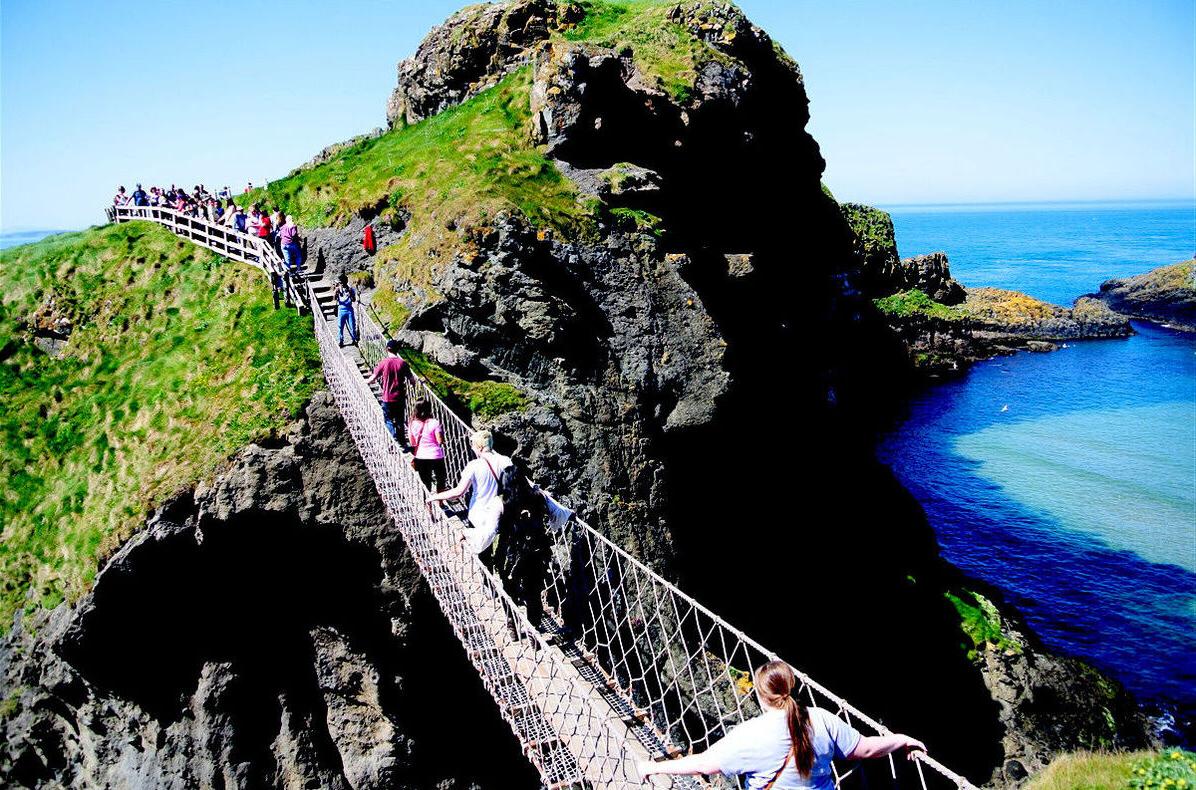访问ors make their way across a rope bridge spanning a gap in a rocky shore area in Ireland